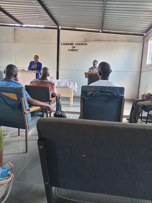 A preacher in a blue suit speaks from a wooden stand beneath the church sign during our church service times in Pretoria