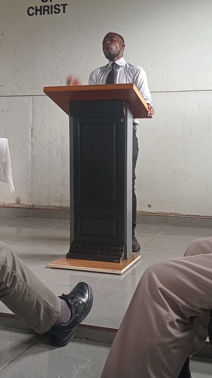 A man in a white shirt and tie teaches from a wooden lectern at our church of Christ in Laudium